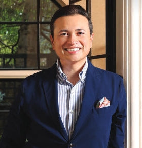 Man wearing a blue blazer and striped shirt standing indoors with a window in the background